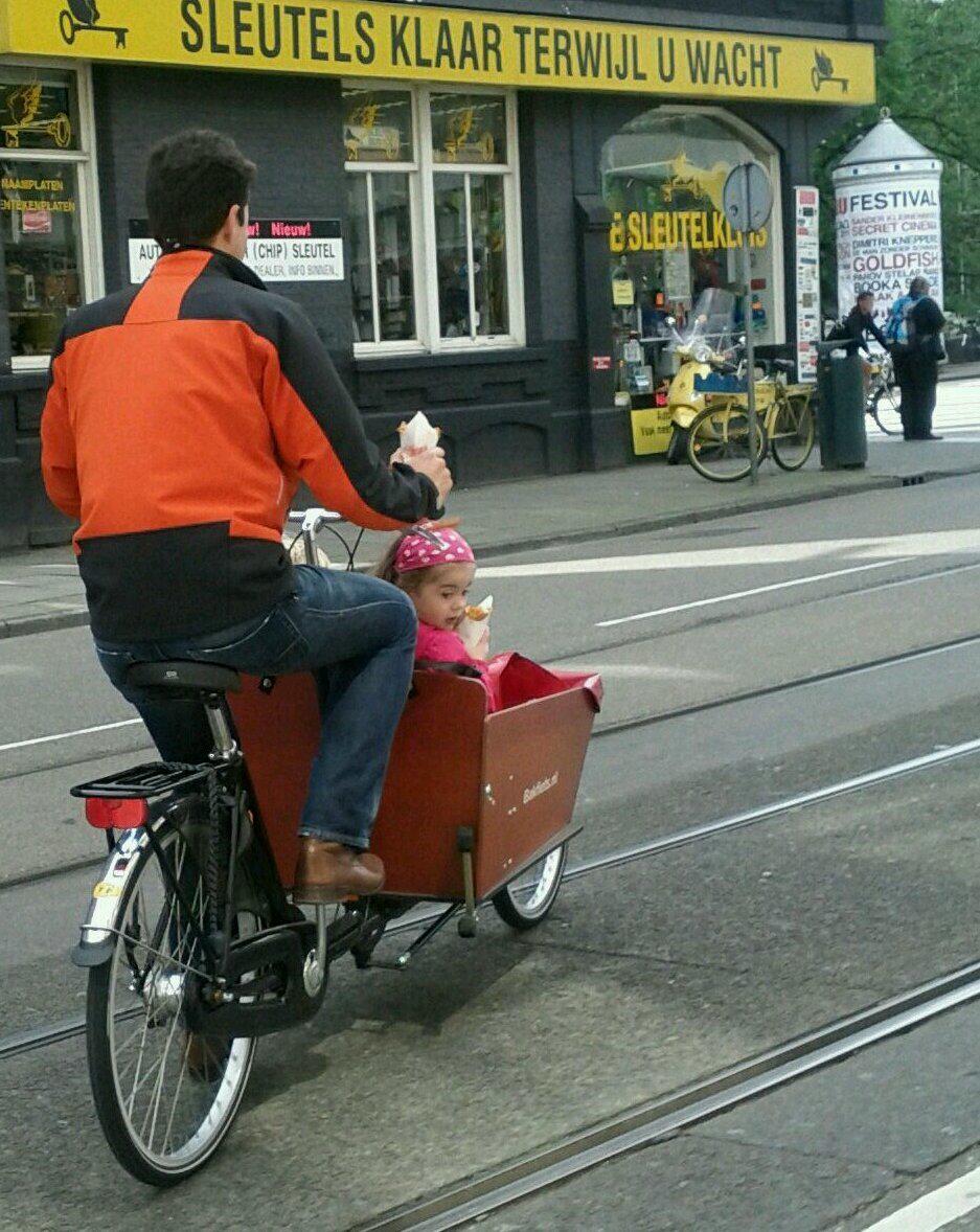 Guy biking with daughter in bakfiets, each holding a cone of frites.